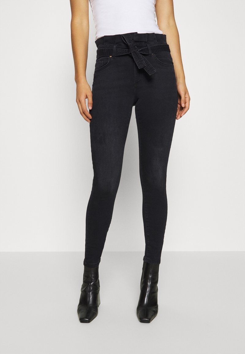 Jean skinny taille haute noir en denim, avec ceinture en tissu nouée, poches avant et texture lisse. Associé à des bottines noires.