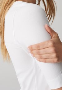 Personne aux longs cheveux portant une chemise blanche à manches longues tenant son bras supérieur avec une main.