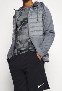 Grå pufferjacka med en strukturerad yta, kombinerad med en svart kamouflagemönstrad t-shirt och svarta shorts med en vit Nike-logotyp.