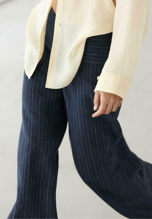 Osoba nosząca luźne granatowe spodnie w prążki i jasno beżową koszulę z długim rękawem, idąca z ręką rozluźnioną wzdłuż ciała.