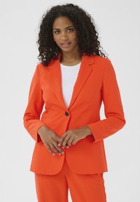 Jasně oranžový přizpůsobený blazer s jednoduchým zapínáním na knoflík, klopovými límci a hladkým materiálem. Nosený přes bílou košili, stylizovaný s ladícími kalhotami.