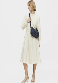 Un vestido midi color crema con cintura fruncida, mangas largas y cuello abotonado. Combinado con un bolso cruzado azul marino y sandalias de cuña blancas.
