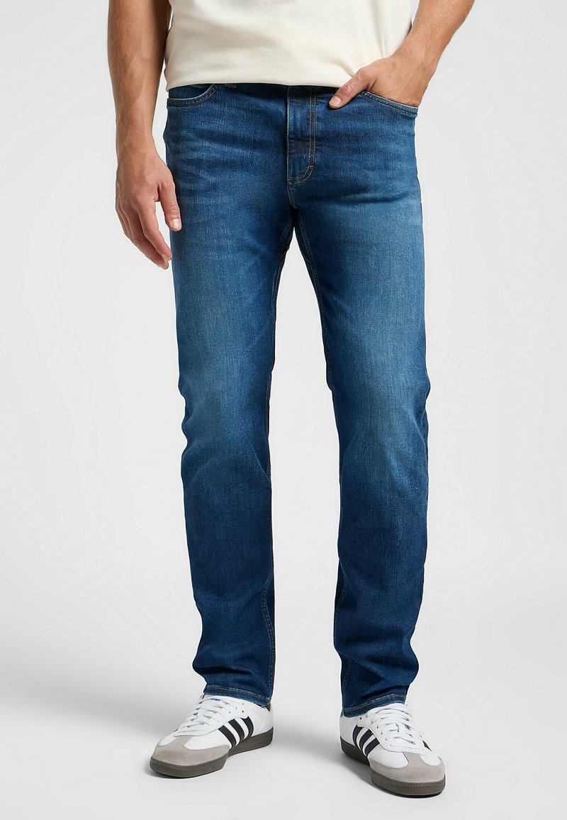 Temno modri jeans s ravnim krojem, petimi žepi, subtilnim bledenjem in kontrastnimi šivi, oblečeni z belimi športnimi čevlji.