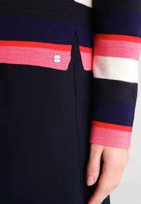 Sweter w paski z dzianiny w odcieniach granatu, bieli i różu, z bocznym rozcięciem i małym logo przy dole. Gładka faktura.