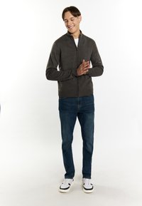 Donkergrijze ritssweater met een hoge kraag, geribbelde zoom en manchetten, gecombineerd met blauwe jeans en witte sneakers, staande op een lichte achtergrond.