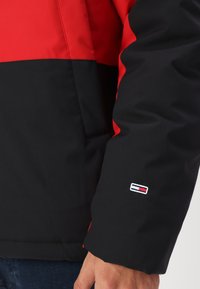 Veste rouge et noire avec une texture lisse et mate. Présente un design en blocs de couleur et un petit logo brodé sur la manche.