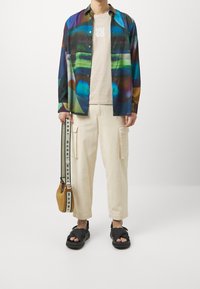 Multicolored patterned shirt, beige t-shirt, cream loose cargo pants, black sandals, and a woven bag with a strap featuring branding.