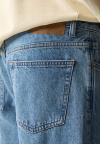 Gros plan sur la poche arrière d'un jean en denim bleu et une étiquette en cuir de la marque sous l'ourlet d'une chemise beige.