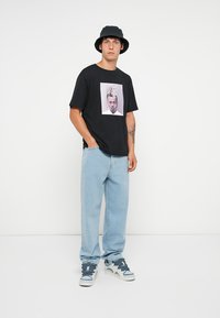 Sort t-shirt med grafisk print, afslappet pasform. Lyseblå jeans og en sort bucket hat. Sneakers med blå og hvide accenter.