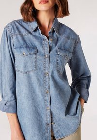 Lysblå denimskjorte med to brystlommer, knappet front, afrundet kant og optrækkede ærmer, med metallic knapper og syninger.
