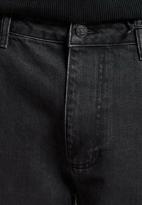 Vaqueros de mezclilla negros con una textura elegante. Cuenta con un cierre frontal de botón, detalles de costura sutiles y un diseño clásico de cinco bolsillos.