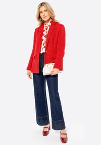 Blazer rosso texture sopra una blusa floreale, abbinato a jeans a gamba larga scuri e ballerine rosse. Una pochette bianca è tenuta in una mano.