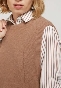 Pull sans manches en tricot marron avec un motif texturé, superposé sur une chemise blanche à rayures horizontales marron et un col chemise.