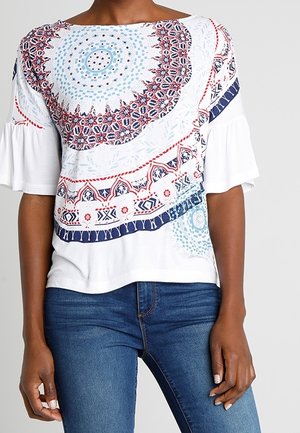 Kobieta ubrana w białą bluzkę z niebiesko-czerwonymi okrągłymi kwiatowymi wzorami i falbanianymi rękawami do łokcia, w połączeniu z niebieskimi dżinsami.