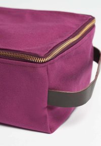 Lila canvasväska med ett guldblixtlås och ett mörkbrunt läderhandtag. Den rektangulära formen har en texturerad yta.