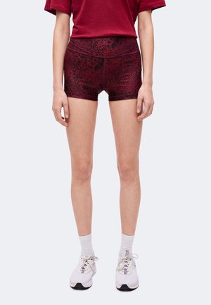 Person iført mørkerøde mønstrede træningsshorts, hvide sokker, hvide sneakers og en mørkerød t-shirt, som står mod en lys baggrund.