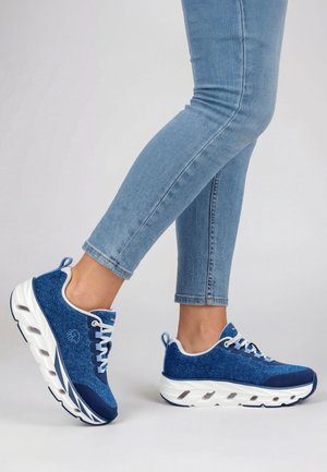 Person trägt blaue Turnschuhe mit dicken weißen Sohlen und hellblaue Jeans vor einem schlichten grauen Hintergrund.
