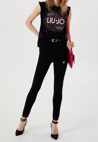 Top de tirantes negro con volantes, con texto rosa; combinado con pantalones negros ajustados con encaje y tacones negros de aguja.