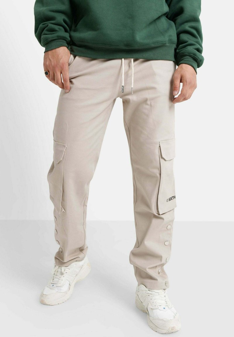 Beige cargobroek met zijzakken, knoopdetails langs de zoom, gedragen met witte sneakers en een groene sweatshirt.