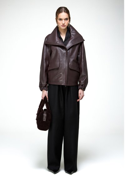 Žena v tmavohnedej nadrozmernej koženej bunde, čiernych širokých nohaviciach, čiernych špicatých topánkach a držiaca hnedú chlpatú kabelku.