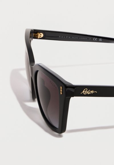 Μαύρα τετράγωνα γυαλιά ηλίου με σκούρους φακούς και χρυσό λογότυπο "Ralph" στο πλευρό, πάνω σε ανοιχτό φόντο.
