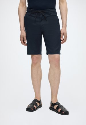 Mann trägt schwarze Shorts mit Kordelzug und schwarze Leder-Sandalen mit offenen Zehen, steht vor einem einfarbigen hellen Hintergrund.