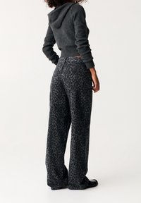 Mujer vestida con un suéter con capucha gris oscuro y pantalones holgados de estampado de leopardo en negro y gris, de pie contra un fondo blanco liso.