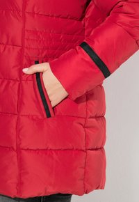 Giacca a vento rossa con cuciture orizzontali, con dettagli neri su tasche e polsini. Texture morbida e design imbottito per calore.