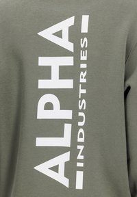 Felpa verde oliva con grandi lettere bianche in maiuscolo che formano la scritta "ALPHA INDUSTRIES" disposta verticalmente lungo il lato, caratterizzata da una morbida trama di cotone.