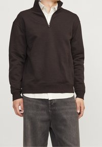 Sudadera marrón con cremallera y cuello alto, textura suave, puños acanalados y un ajuste relajado, usada sobre una camisa blanca de cuello.