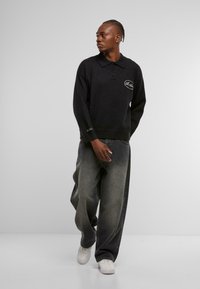 Schwarzes Langarm-Polo mit drei Knöpfen und gesticktem Logo. Getragen mit locker sitzenden, allmählich ausgewaschenen grauen Jeans und weißen Sneakers.