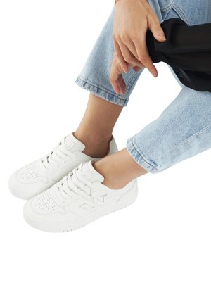 Baskets blanches en cuir avec des détails perforés, fermeture à lacets et semelle texturée, associées à un jean en denim retroussé.