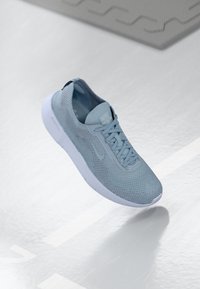 Vaaleansininen juoksukenkä, jossa on hengittävä verkkokangas yläosa, tasaiset nauhat ja pehmeä valkoinen pohja. Ominaisuuksiin kuuluu Nike-logo kielekkeessä ja sivussa.