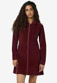 Burgundy kordflöjlsklänning med långa ärmar, dragkedja fram och bröstficka. Har vertikal ribbtextur och en utsvängd nederdel.