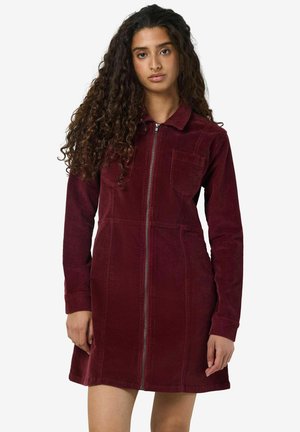 Bordeaux corduroy jurk met lange mouwen, ritssluiting aan de voorkant en een borstzak. Heeft een verticale ribbelstructuur en een uitlopende zoom.