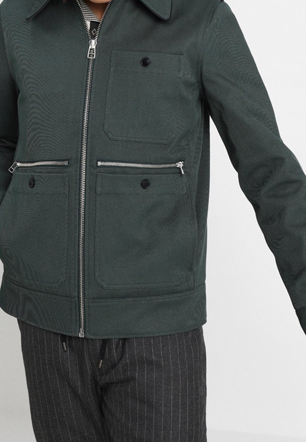 Dunkelgrüne Jacke aus strukturiertem Stoff. Verfügt über einen Reißverschluss vorne, zwei Seitentaschen und eine Brusttasche mit Knopfakzenten.