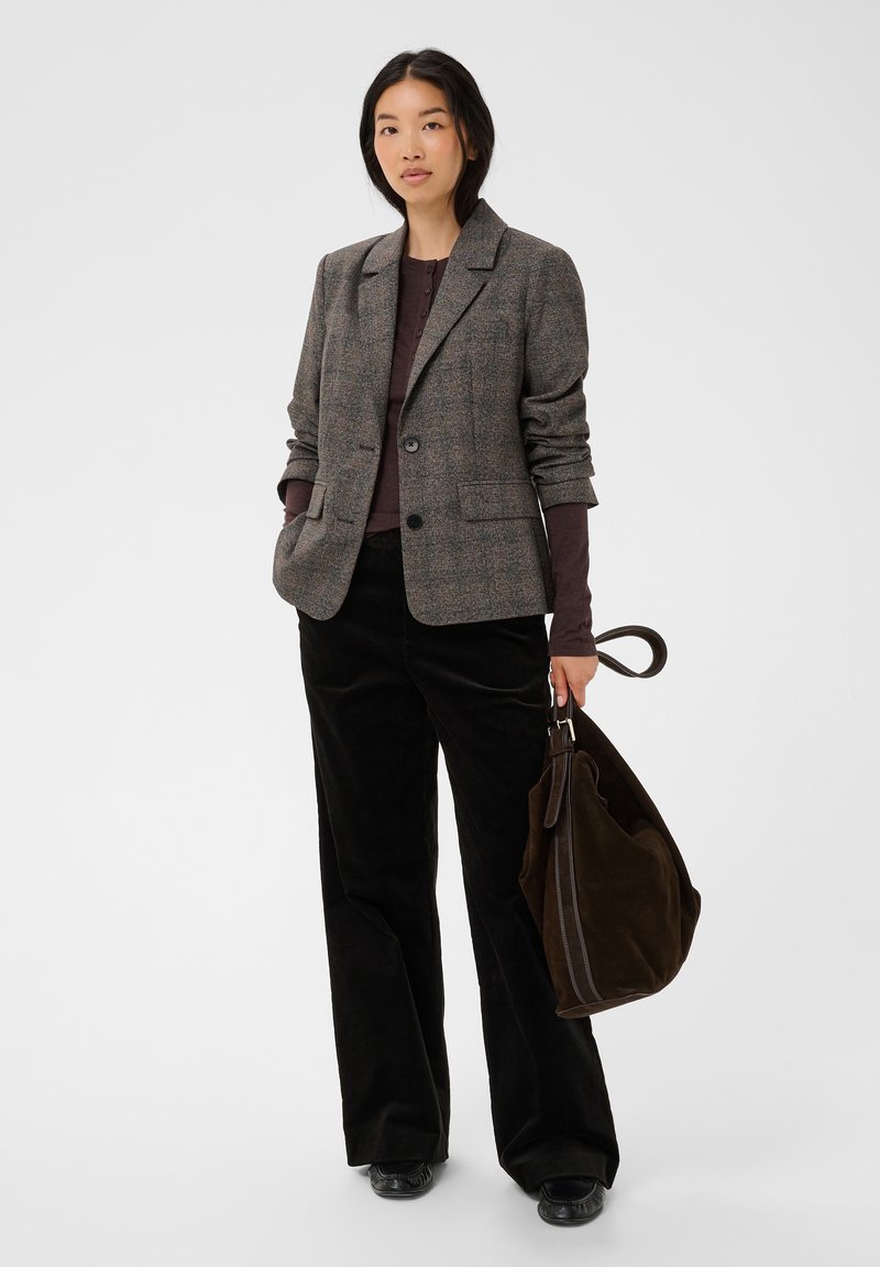 Femme portant un blazer à carreaux gris, un haut à manches longues foncé, un pantalon large noir, tenant un grand sac en daim marron avec une bandoulière.