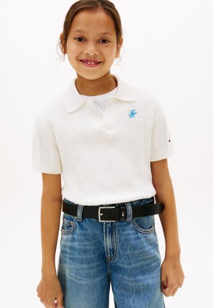 Niña sonriente con un suéter tipo polo blanco de manga corta y textura, metido dentro de jeans azules con un cinturón negro, sobre un fondo liso.