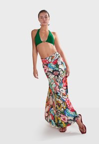 Groene halter bikini-top gecombineerd met een lange, bloemige maxi-jurk met levendige kleuren en een vloeiende pasvorm. Platte rode sandalen maken de look compleet.