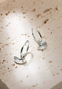 Boucles d'oreilles en argent avec un pendentif en forme de éventail présentant des détails plissés, exposées sur une surface texturée de couleur claire.