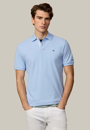 Joven con camiseta polo azul claro con pequeño logo y pantalones blancos, de pie con las manos en los bolsillos frente a un fondo beige.