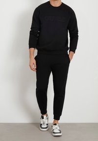 Man in een zwarte GUESS-hoodie, zwarte joggingsbroek en witte sneakers met zwarte accenten, staand met één hand in de zak.