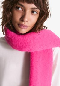 Roza volnena šala z mehko, puhasto teksturo, sproščeno ovita okoli vratu in dopolnjuje svetlo oblečeno majico.