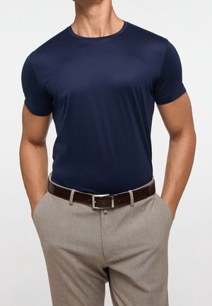 Uomo che indossa una camicia blu navy a maniche corte infilata in pantaloni beige con una cintura marrone scuro, mani nelle tasche, su sfondo semplice.
