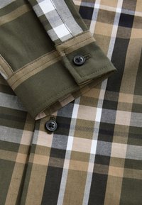 Chemise à carreaux aux nuances de vert olive, beige et noir. Fabriquée en coton avec une texture lisse. Comprend deux boutons noirs sur le poignet.