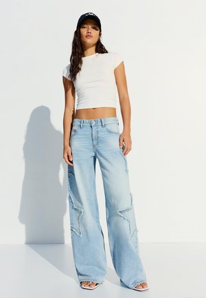 Kobieta ubrana w biały krótki top, jasnoniebieskie szerokie jeansy z naszywkami w kształcie gwiazd, czarną czapkę i białe sandały na jednolitym białym tle.