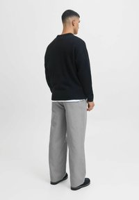 Pulover negru tricotat, pantaloni gri cu croială largă și pantofi negri. Modelul este văzut din spate pe un fundal alb simplu.