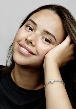 Braccialetto charm in argento con un pendente a forma di cuore, caratterizzato da una superficie liscia. Indossato su un polso, con una maglietta nera visibile.