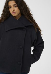 Schwarzer Wollmischmantel mit weitem Kragen, großen Knöpfen und kastiger Silhouette. Weiche Textur, präsentiert an einem Modell vor neutralem Hintergrund.