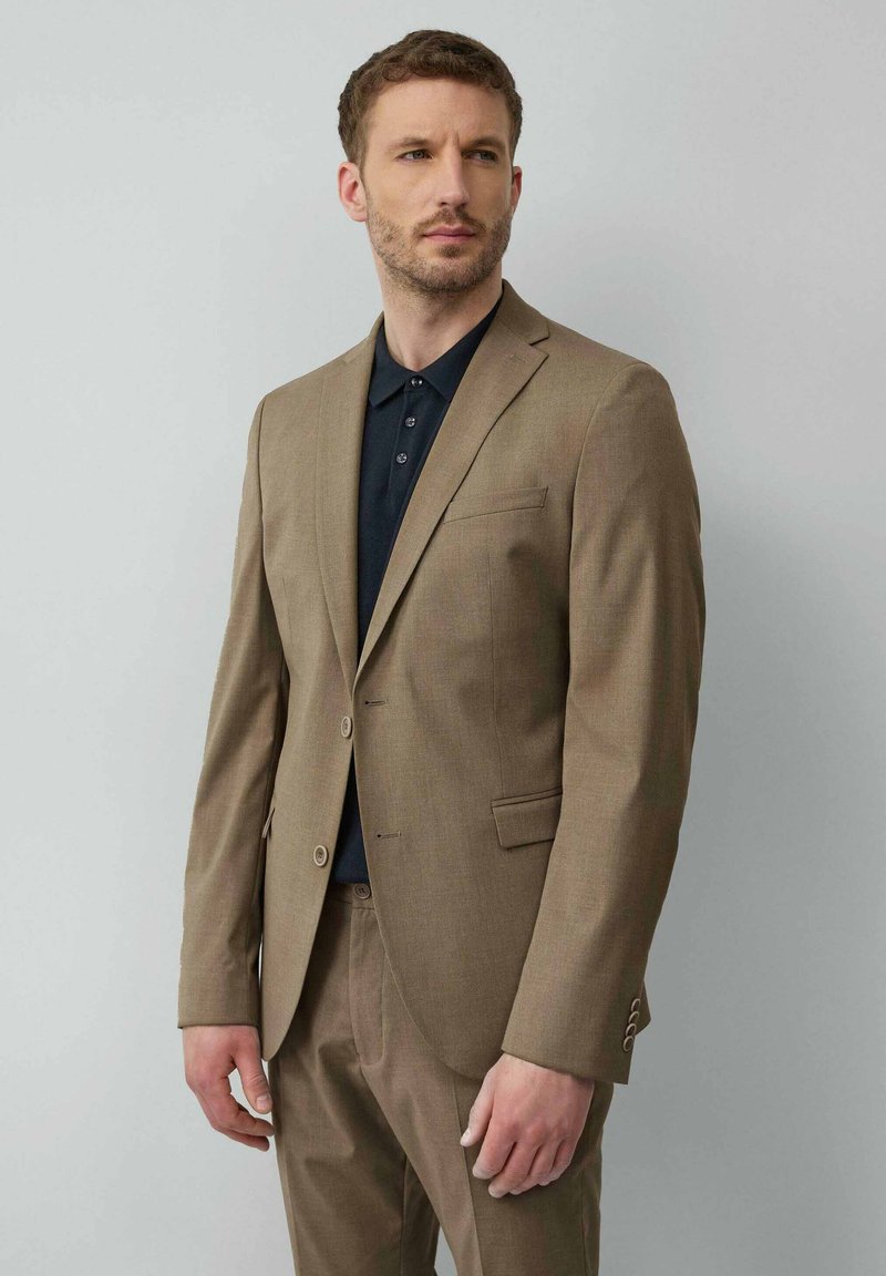 Lichtbruine pak met een getailleerd ontwerp, twee knopen en een borstzak, gecombineerd met een donker polohemd eronder. Duidelijke textuur.